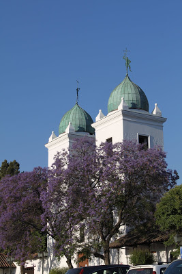 White building with two green steeples and trees with purple flowers in the foreground