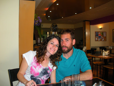 woman on left leaning into man on right, sitting at a table with glasses and dark restaurant in the background