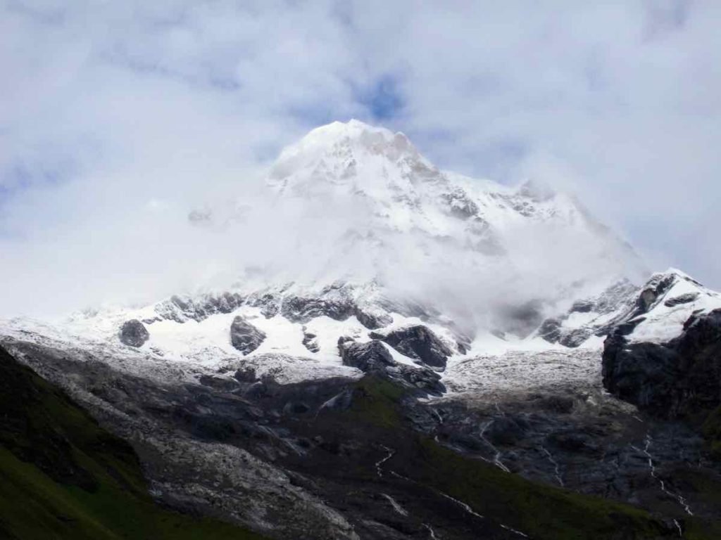 mountain peak with snow, lots of cloudy but a bit of blue sky peaking though, dark river bed in front
