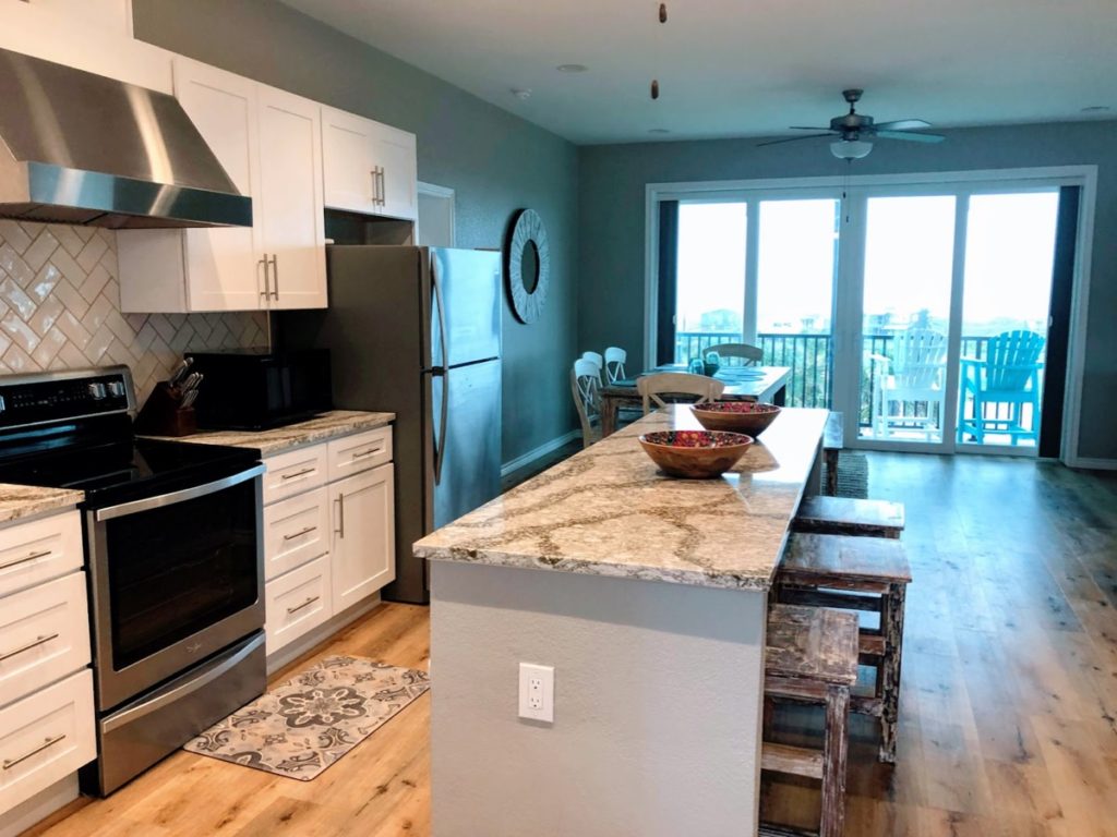 Inside of vacation rental in Port Aransas, you see the kitchen with island, dining table, and a porch with chairs facing the ocean.